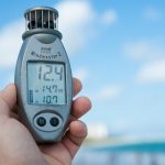 Close-up of a hand holding Windmaster 2 anemometer against a clear blue sky and beach backdrop.
