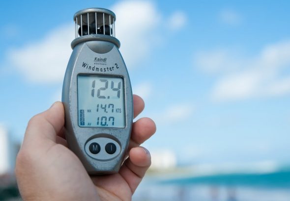 Close-up of a hand holding Windmaster 2 anemometer against a clear blue sky and beach backdrop.
